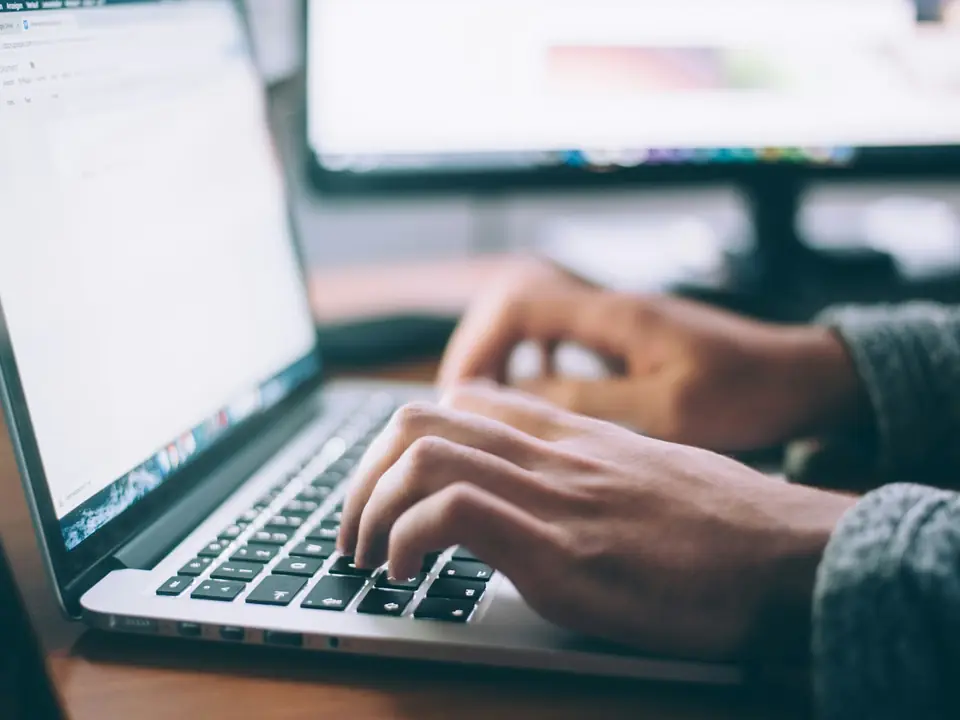 Manos escribiendo en el teclado de un portátil sobre una mesa de trabajo con pantallas al fondo